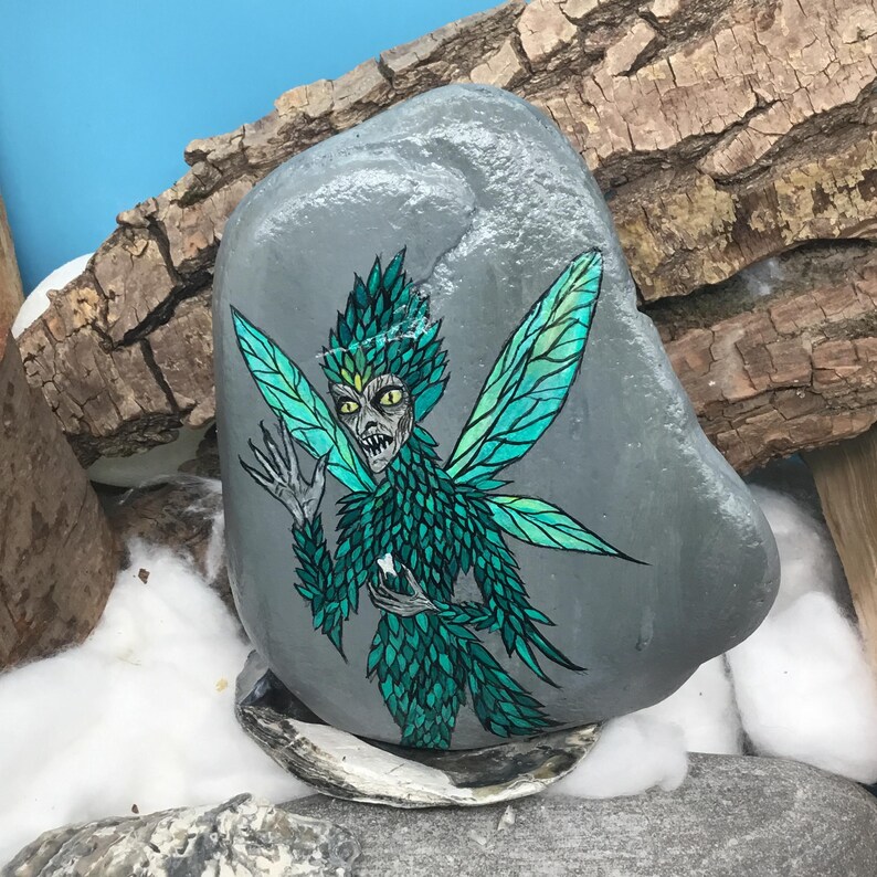 May include: A hand-painted, gray stone features a detailed illustration of a fairy. The fairy has green and teal wings, a matching feathered outfit, and a stern expression. The artwork is set against a backdrop of wood and cotton.