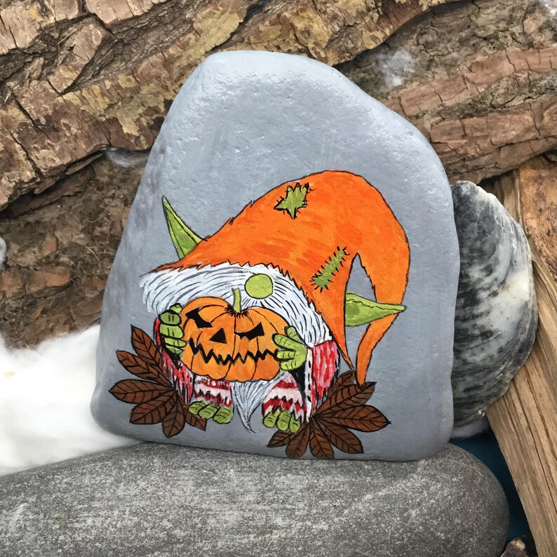 May include: A hand-painted, gray rock featuring a Halloween gnome holding a carved pumpkin. The gnome wears an orange hat and green ears. The pumpkin is orange with a carved face. Brown leaves and white beard complete the design.
