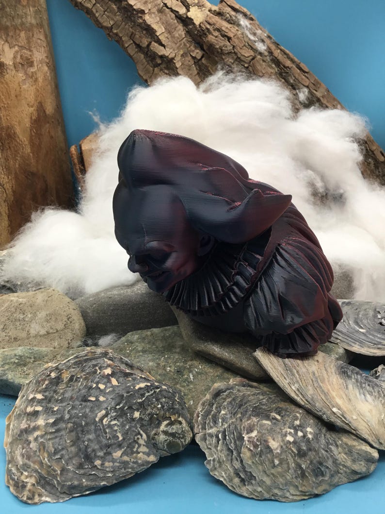 May include: A dark burgundy and black sculpture of a figure with a ruffled collar and a large hat. The sculpture is resting on a bed of gray stones and oyster shells, with a backdrop of white fluff and wood.