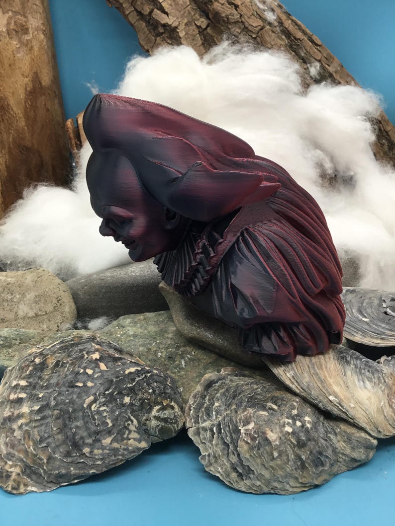 May include: A bust sculpture of a person with a large hat and ruffled collar, in shades of burgundy and black. The sculpture is resting on a bed of rocks and oyster shells, with a backdrop of cotton and wood.