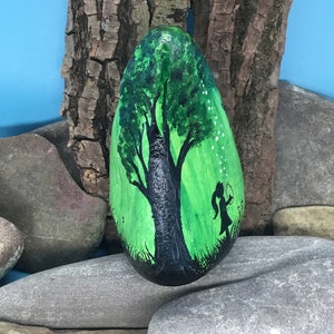 K&ouml;nnte beinhalten: Handbemalter Stein mit der Silhouette eines Baumes mit gr&uuml;nen Bl&auml;ttern und einem M&auml;dchen, das L&ouml;wenzahnsamen in ein gr&uuml;nes Feld bl&auml;st.