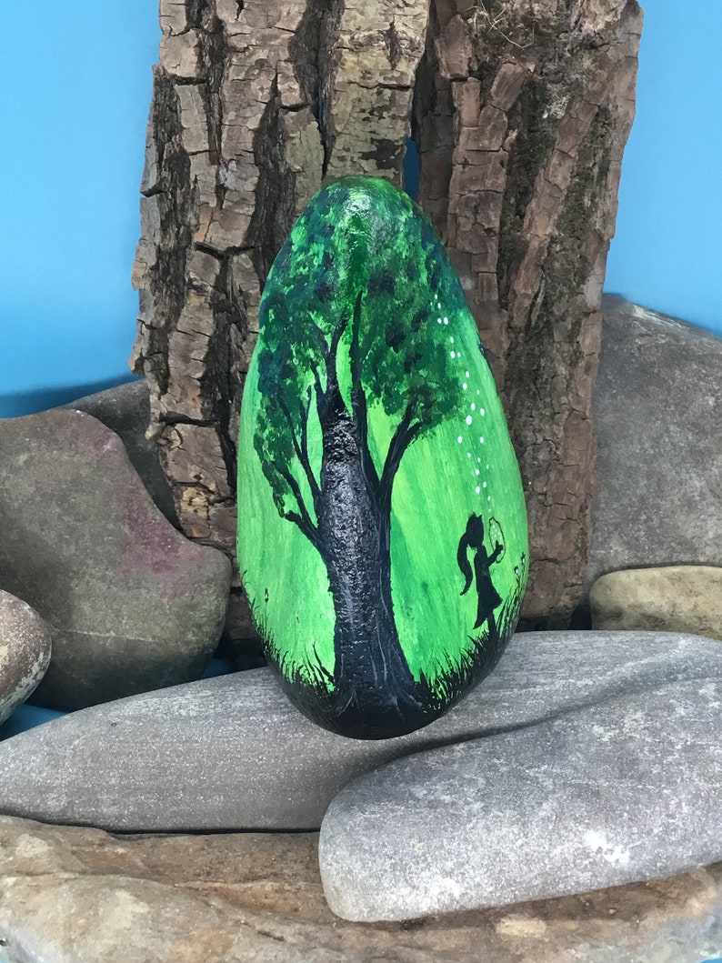 K&ouml;nnte beinhalten: Handbemalter Stein mit der Silhouette eines Baumes mit gr&uuml;nen Bl&auml;ttern und einem M&auml;dchen, das L&ouml;wenzahnsamen in ein gr&uuml;nes Feld bl&auml;st.