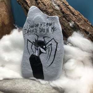 May include: A grey painted rock featuring a black ink illustration of a slender, multi-limbed creature with a sharp grin. Above the figure, the text reads: I DON'T LIVE IN DARKNESS DARKNESS LIVES IN ME. The rock has an irregular, jagged top edge.