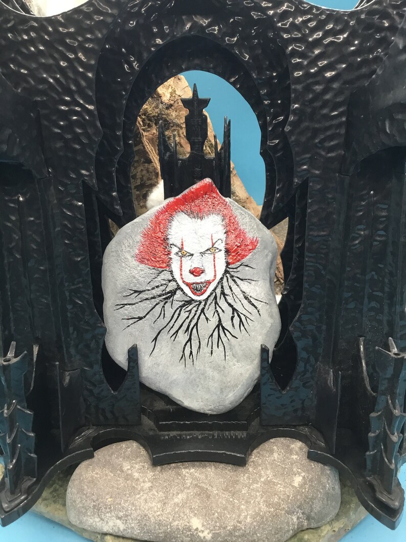 May include: A hand-painted rock with a depiction of Pennywise the Dancing Clown from the movie "IT". The rock is placed in a black plastic frame with a gothic archway design. The background is a blue sky with a silhouette of a castle.