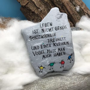 May include: A light gray painted stone featuring black hand-lettered German text that reads LEBEN IST NICHT GENUG SONNENSCHEIN FREIHEIT UND EINEN KLEINEN VOGEL MUSS MAN AUCH HABEN. Below the text are four small, colorful bird illustrations in yellow, green, red, and blue.