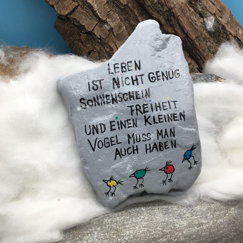 May include: A light gray painted stone featuring black hand-lettered German text that reads LEBEN IST NICHT GENUG SONNENSCHEIN FREIHEIT UND EINEN KLEINEN VOGEL MUSS MAN AUCH HABEN. Below the text are four small, colorful bird illustrations in yellow, green, red, and blue.