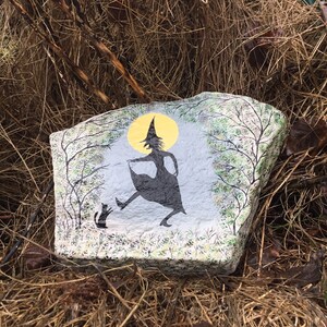 May include: A painted rock with a silhouette of a witch and a cat in front of a full moon. The witch is wearing a pointed hat and a long dress. The rock is surrounded by dry grass.