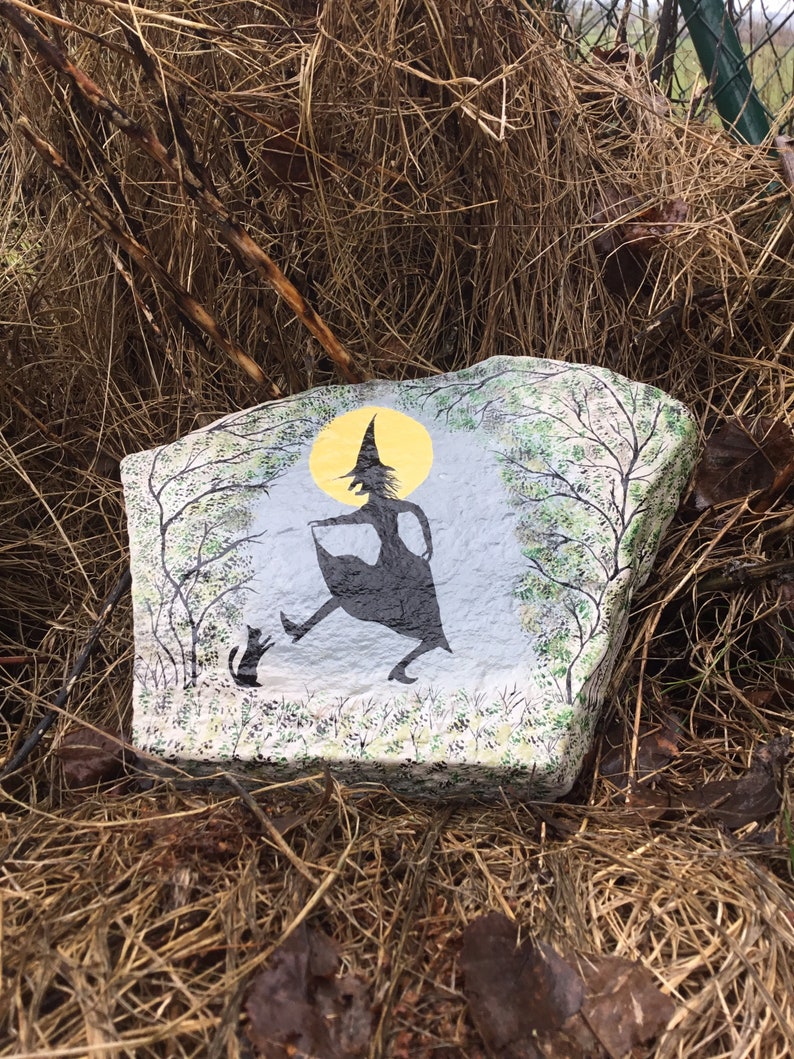 May include: A painted rock with a silhouette of a witch and a cat in front of a full moon. The witch is wearing a pointed hat and a long dress. The rock is surrounded by dry grass.