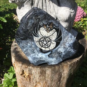 May include: A hand-painted rock featuring a bat with a pentagram design. The bat has black wings and a tan face, set against a white and black background. The rock is dark gray and sits on a wooden stump, with greenery in the background.