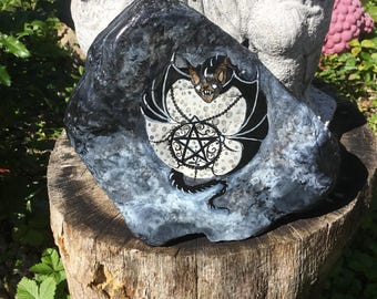The Gothic bat embraces the moon with an amulet and pentagram, a unique, hand-painted piece immortalized on a natural stone in comic style.