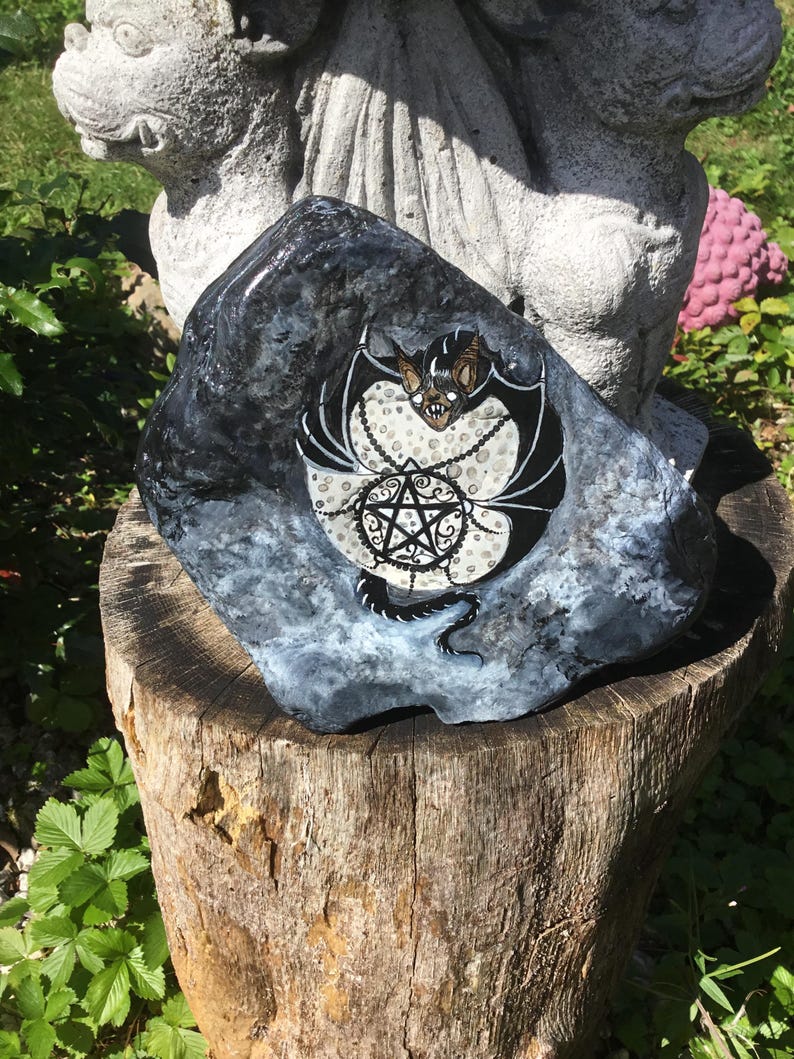 May include: A hand-painted rock featuring a bat with a pentagram design. The bat has black wings and a tan face, set against a white and black background. The rock is dark gray and sits on a wooden stump, with greenery in the background.