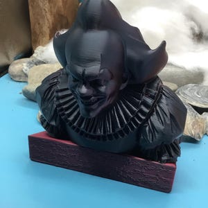 May include: A black bust of a menacing clown with a ruffled collar and a sinister grin. The sculpture is mounted on a rectangular, maroon base. The clown's hair is styled in a large, pointed shape.