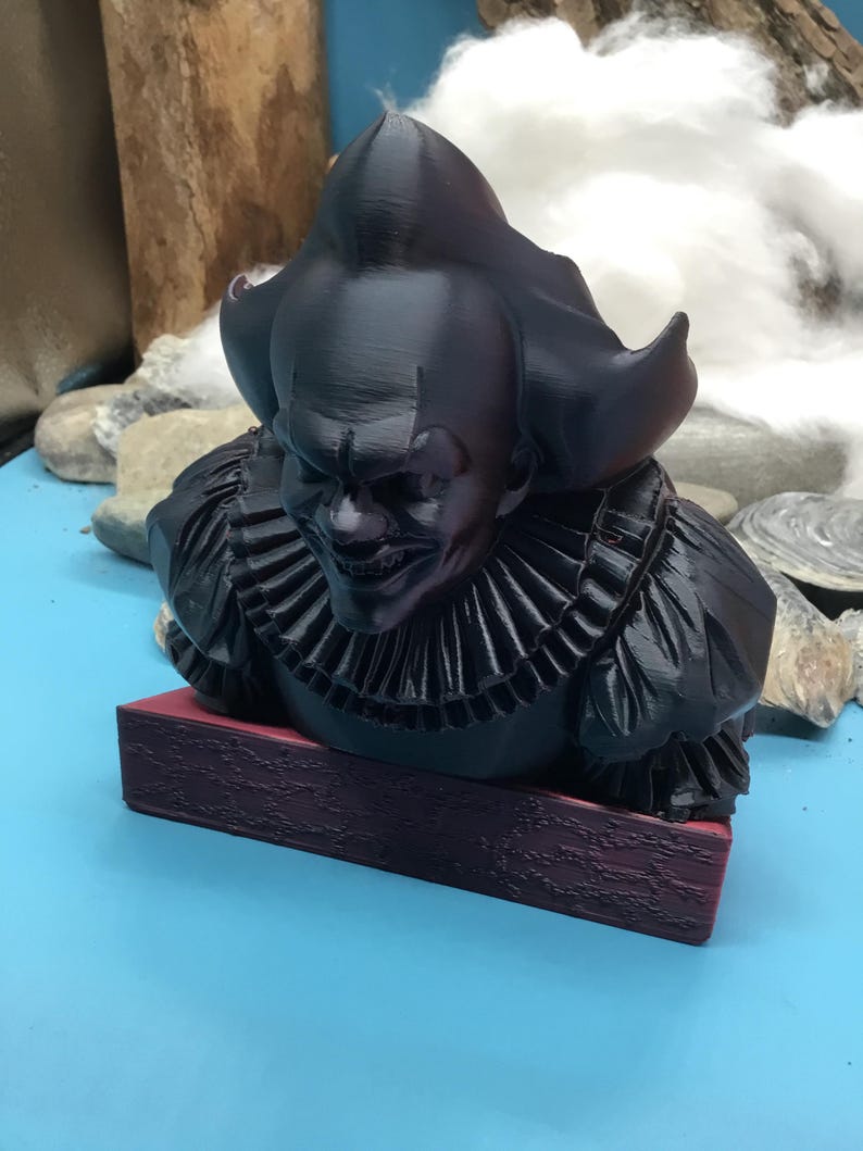 May include: A black bust of a menacing clown with a ruffled collar and a sinister grin. The sculpture is mounted on a rectangular, maroon base. The clown's hair is styled in a large, pointed shape.