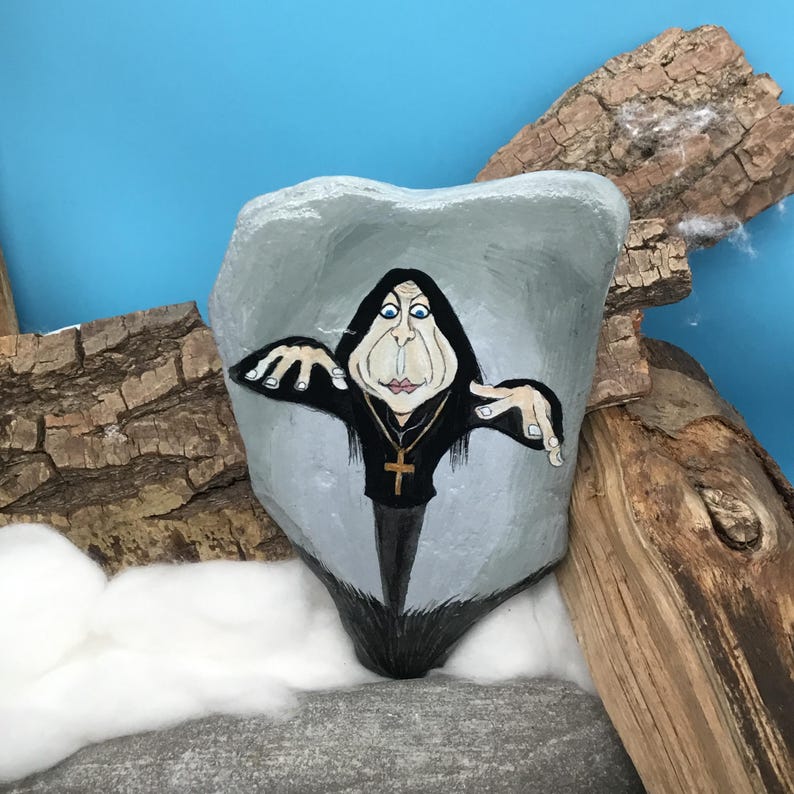 May include: A hand-painted rock depicts a cartoon figure in a black robe and gold cross necklace. The figure has a pale face with blue eyes, set against a grey background. The rock is surrounded by wood and cotton wool.