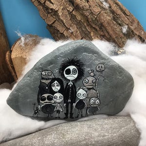 Puede incluir: Una piedra gris lisa con una detallada ilustraci&oacute;n pintada a mano de un grupo de personajes estilizados de inspiraci&oacute;n g&oacute;tica. La obra muestra una figura central alta con cabello negro desordenado rodeada de varias figuras m&aacute;s peque&ntilde;as.