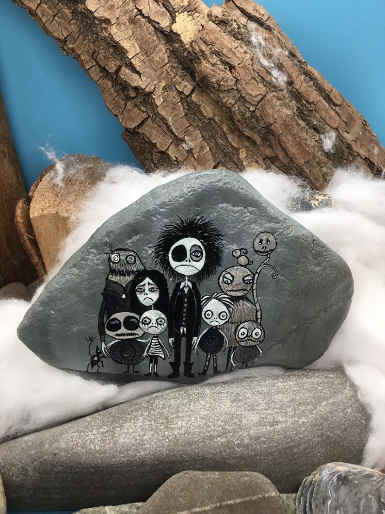 Puede incluir: Una piedra gris lisa con una detallada ilustraci&oacute;n pintada a mano de un grupo de personajes estilizados de inspiraci&oacute;n g&oacute;tica. La obra muestra una figura central alta con cabello negro desordenado rodeada de varias figuras m&aacute;s peque&ntilde;as.