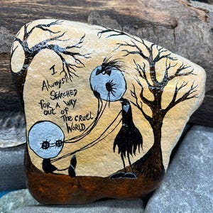 Peut inclure: Une pierre peinte &agrave; la main repr&eacute;sentant une sc&egrave;ne fantaisiste avec deux personnages ronds et un personnage grand et mince sous des arbres d&eacute;nud&eacute;s. Le texte "I always searched for a way out of the cruel world" est &eacute;crit sur la pierre.