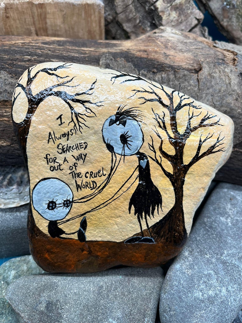 Peut inclure: Une pierre peinte &agrave; la main repr&eacute;sentant une sc&egrave;ne fantaisiste avec deux personnages ronds et un personnage grand et mince sous des arbres d&eacute;nud&eacute;s. Le texte "I always searched for a way out of the cruel world" est &eacute;crit sur la pierre.