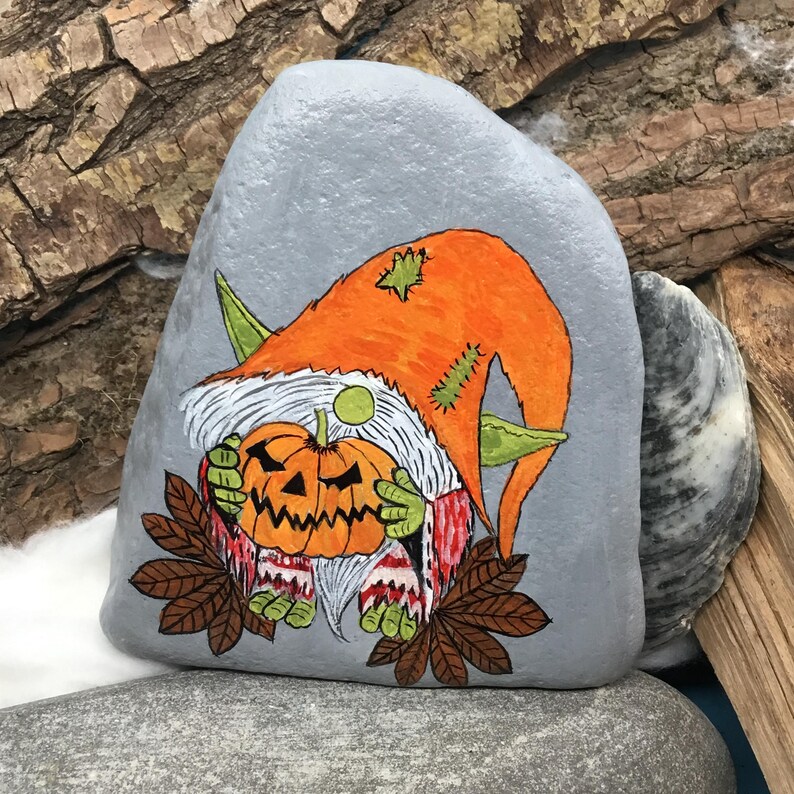 May include: A hand-painted, gray stone features a Halloween gnome holding a carved pumpkin. The gnome wears an orange hat with green accents and a white beard. The pumpkin is orange with a carved face. Brown leaves are at the bottom of the stone.