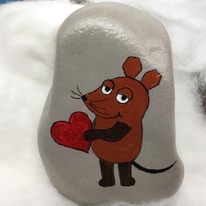 Pode incluir: Uma pedra cinza pintada à mão apresenta um rato de desenho animado segurando um coração vermelho. O rato é marrom com detalhes em preto e uma cauda longa e curvada. O fundo é um material branco e macio.