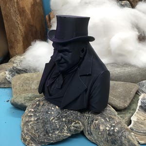 May include: A dark navy bust of a man in a top hat and suit jacket. The sculpture is placed on a large, textured oyster shell. The background features rocks, wood, and white fluffy material, set against a blue surface.