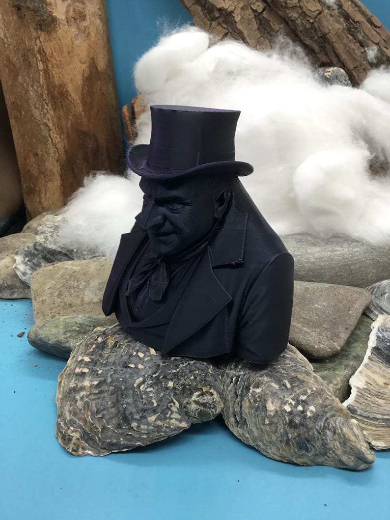 May include: A dark navy bust of a man in a top hat and suit jacket. The sculpture is placed on a large, textured oyster shell. The background features rocks, wood, and white fluffy material, set against a blue surface.