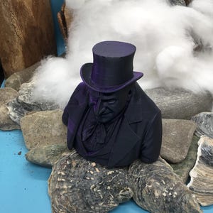 May include: A dark purple bust of a man in a top hat and suit. The bust is set amongst rocks, oyster shells, and cotton. The man's face is detailed, with a serious expression. The bust is a decorative item.