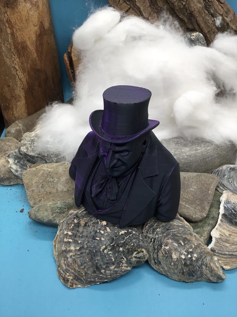 May include: A dark purple bust of a man in a top hat and suit. The bust is set amongst rocks, oyster shells, and cotton. The man's face is detailed, with a serious expression. The bust is a decorative item.