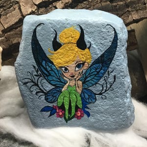 May include: Hand-painted rock art featuring a fairy with blue wings, green dress, and yellow hair. The fairy is sitting on a flower with pink petals. The background is a light blue color.