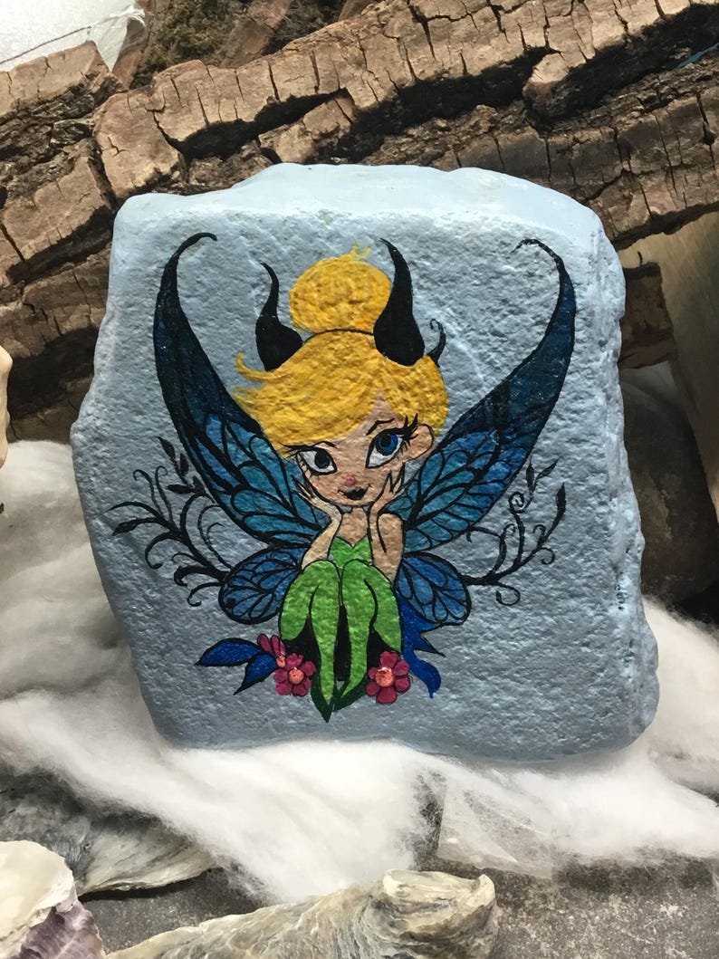 May include: Hand-painted rock art featuring a fairy with blue wings, green dress, and yellow hair. The fairy is sitting on a flower with pink petals. The background is a light blue color.