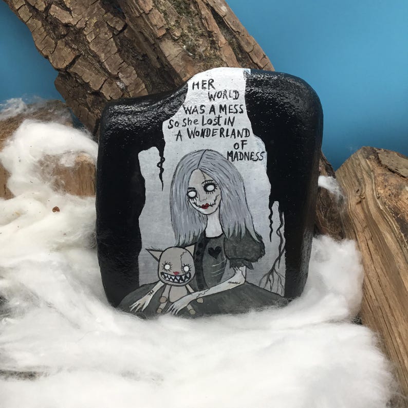 May include: A hand-painted, black rock featuring a gothic illustration. The artwork depicts a pale-skinned woman with long gray hair and a doll, with the text "Her world was a mess so she lost in a wonderland of madness."