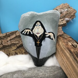 May include: A painted rock depicts a cartoon figure in black robes, wearing a gold cross necklace. The figure has a pale face with blue eyes, set against a grey background. The rock is displayed on white fluff and a blue backdrop, with wood pieces.