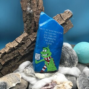 Pode incluir: Uma pedra azul pintada &agrave; m&atilde;o com uma personagem de desenho animado verde com um cachecol vermelho e branco. A pedra tem texto em alem&atilde;o e &eacute; rodeada por conchas, pedras e elementos decorativos.