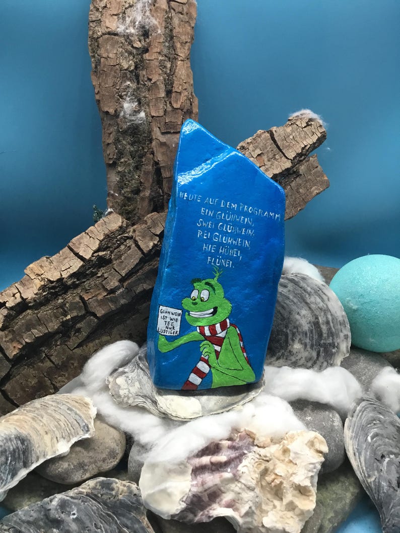 Pode incluir: Uma pedra azul pintada &agrave; m&atilde;o com uma personagem de desenho animado verde com um cachecol vermelho e branco. A pedra tem texto em alem&atilde;o e &eacute; rodeada por conchas, pedras e elementos decorativos.