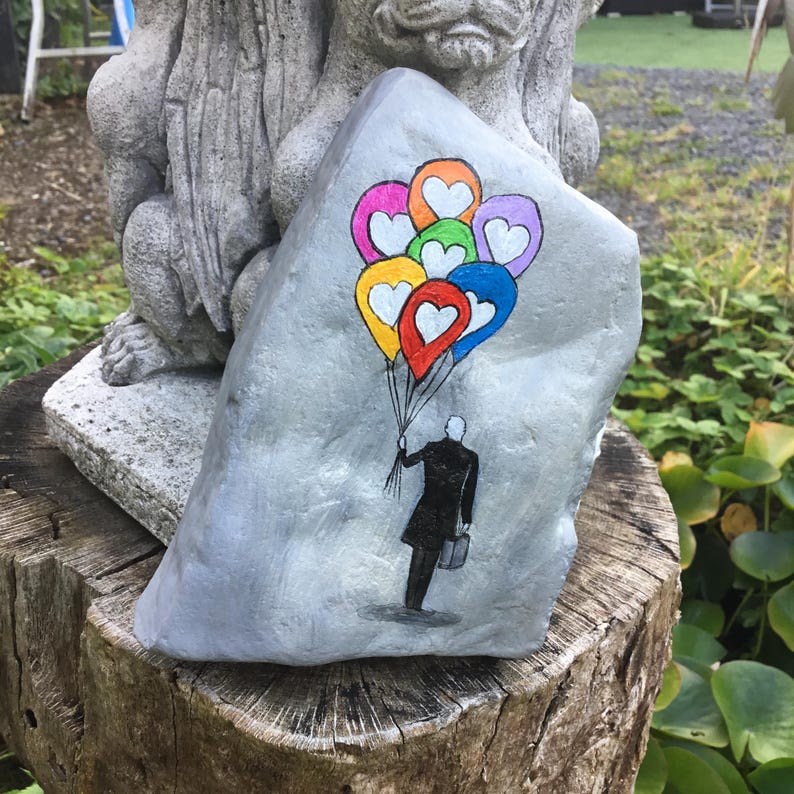 May include: A hand-painted, gray rock features a person in a black coat holding colorful heart-shaped balloons. The balloons are in shades of red, orange, yellow, green, blue, pink, and purple. The rock is displayed on a wooden stump.