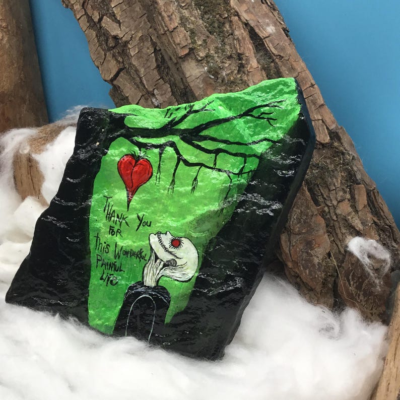 May include: A painted rock with a green and black design. The rock features a red heart, a stylized figure, and the text "Thank You For This Wonderful Painful Life."