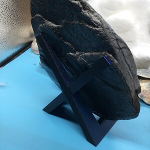May include: A dark grey, textured rock displayed on a dark blue, angled stand. The rock has a rough surface with visible ridges and crevices. The stand is a simple, geometric design, providing support for the stone. The background is a light blue surface.