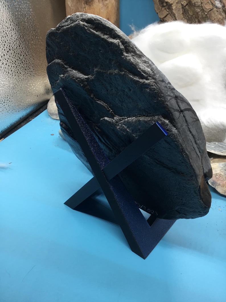 May include: A dark grey, textured rock displayed on a dark blue, angled stand. The rock has a rough surface with visible ridges and crevices. The stand is a simple, geometric design, providing support for the stone. The background is a light blue surface.