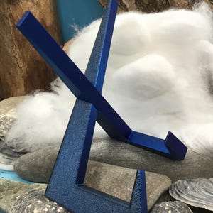 May include: A blue metal display stand with a textured finish. The stand is designed to hold an item and is supported by a base. The stand is made of metal and has a modern design.
