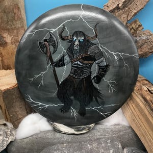 May include: A circular, grey decorative item depicting a minotaur warrior with an axe, set against white lightning bolts. The minotaur has black fur, horns, and blue eyes. The artwork is on a stone-like surface.