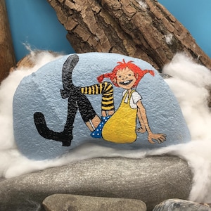 May include: A light blue painted rock featuring a hand-painted illustration of a character with red hair in pigtails, wearing a yellow dress, blue polka-dot shorts, and black boots with yellow and black striped stockings, sitting in a relaxed pose.