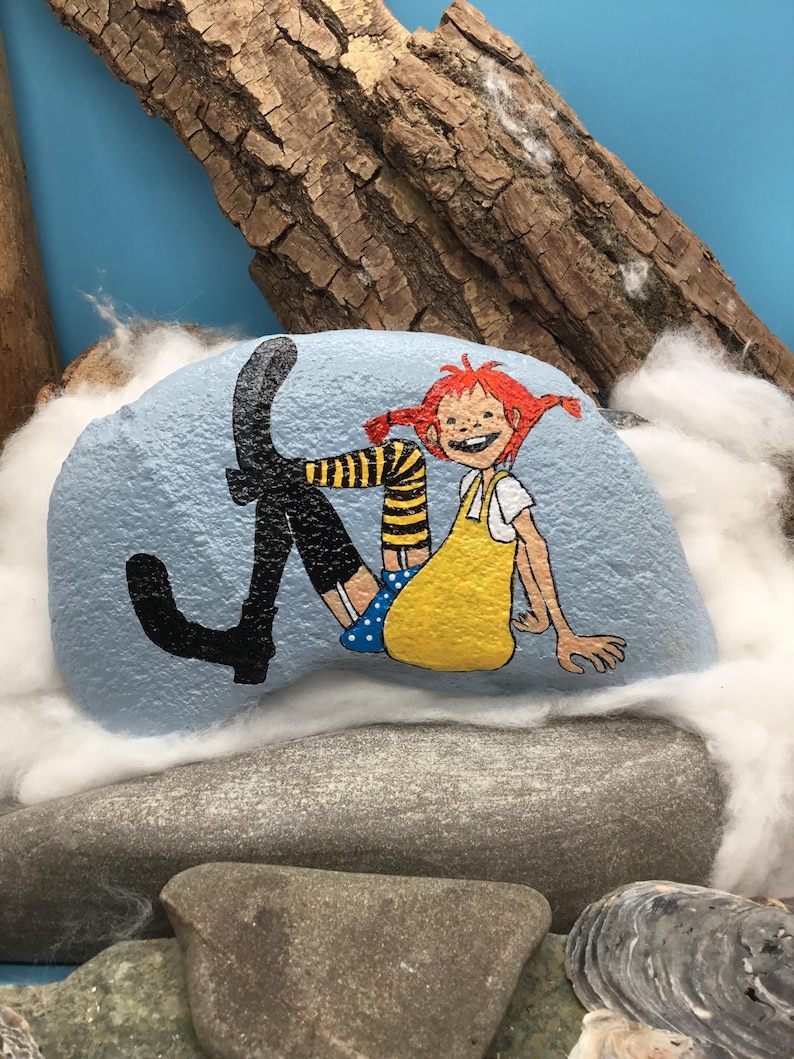 May include: A light blue painted rock featuring a hand-painted illustration of a character with red hair in pigtails, wearing a yellow dress, blue polka-dot shorts, and black boots with yellow and black striped stockings, sitting in a relaxed pose.