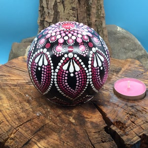 Op de afbeelding: Een zwarte bol met een roze en wit mandala-ontwerp erop geschilderd. De bol ligt op een boomstronk met een roze kaars in een houder ernaast.