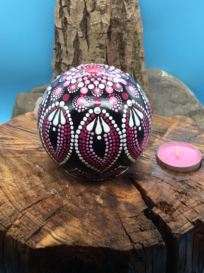 Op de afbeelding: Een zwarte bol met een roze en wit mandala-ontwerp erop geschilderd. De bol ligt op een boomstronk met een roze kaars in een houder ernaast.