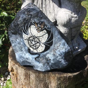 May include: A hand-painted rock featuring a bat design with a pentagram. The bat has black wings and a brown face, set against a white and black background. The rock is gray and sits on a wooden stump, with a statue in the background.