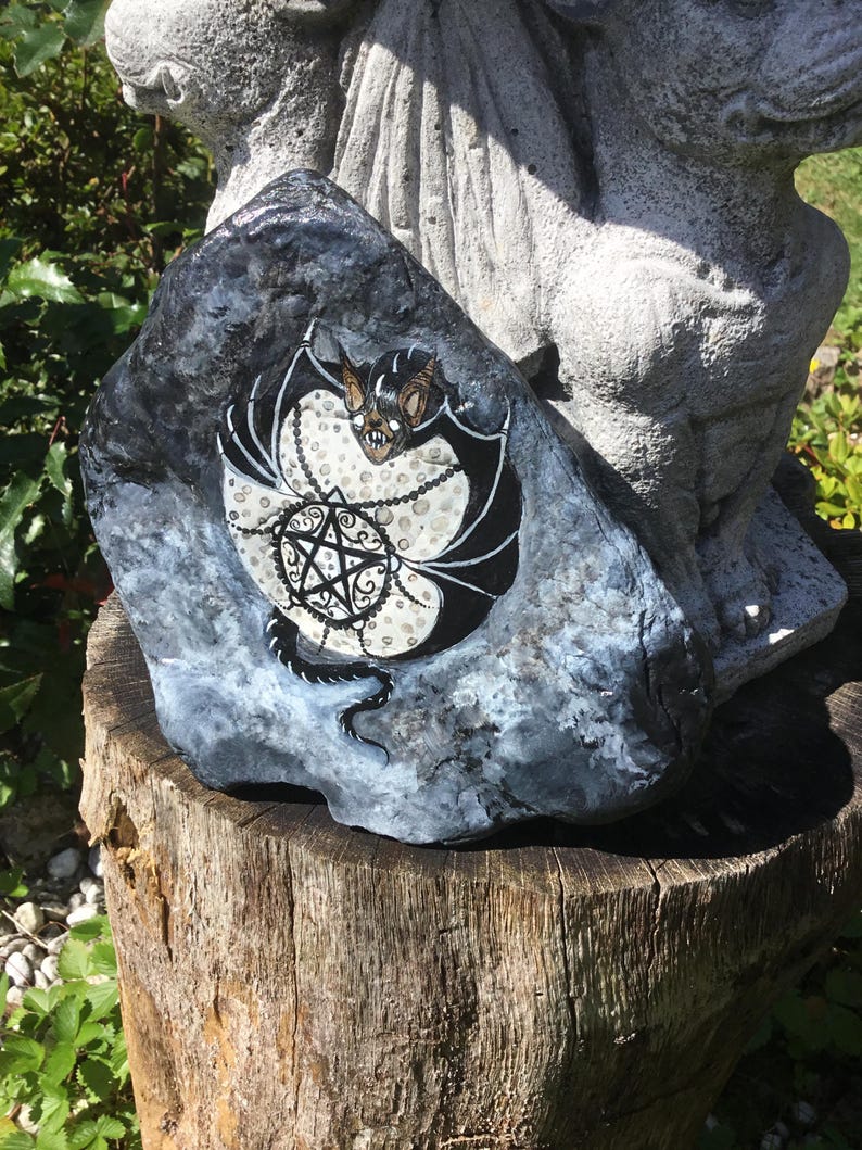 May include: A hand-painted rock featuring a bat design with a pentagram. The bat has black wings and a brown face, set against a white and black background. The rock is gray and sits on a wooden stump, with a statue in the background.