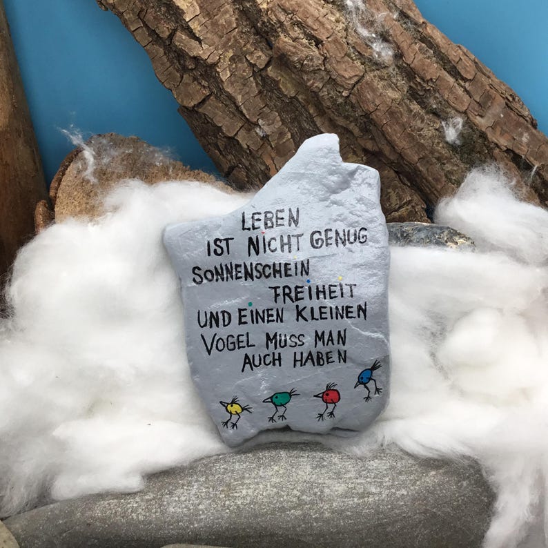 May include: A light gray painted rock featuring the black text LEBEN IST NICHT GENUG SONNENSCHEIN FREIHEIT UND EINEN KLEINEN VOGEL MUSS MAN AUCH HABEN. Below the text are four small hand-drawn birds in yellow, green, red, and blue colors.