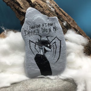 May include: A grey painted rock featuring a black ink illustration of a stylized figure with long, spindly limbs and a white eye. The text "I DON'T LIVE IN DARKNESS DARKNESS LIVES IN ME" is written in black, jagged, hand-lettered script above the figure.
