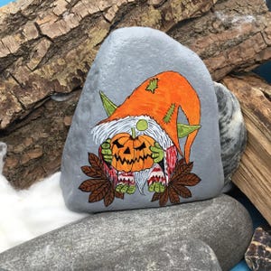 May include: A hand-painted, gray stone features a Halloween gnome holding a carved pumpkin. The gnome wears an orange hat and green pointed ears. The pumpkin is orange with a carved face. Brown leaves and white beard complete the design.