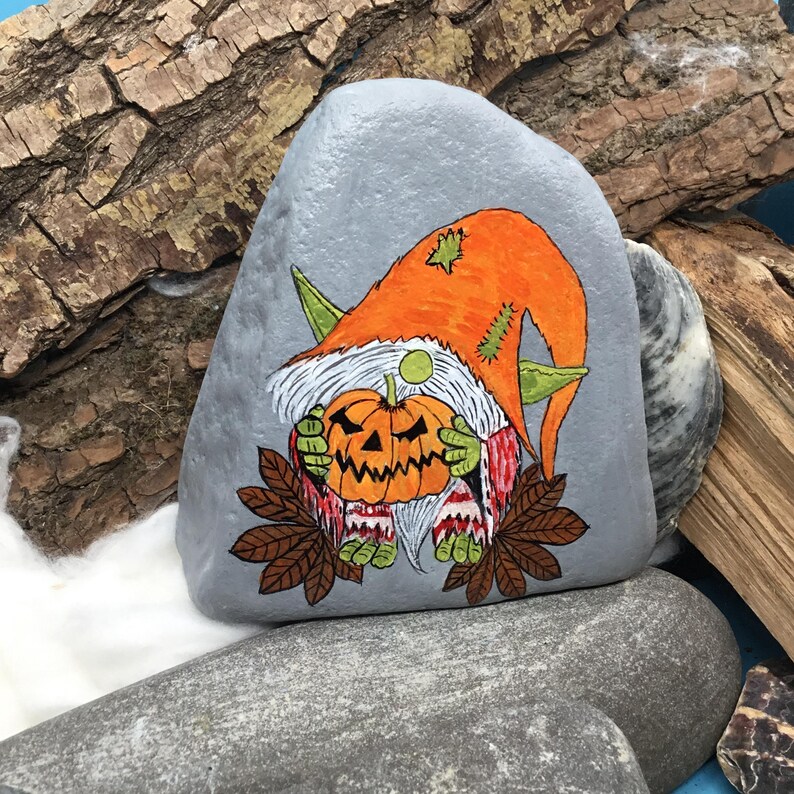May include: A hand-painted, gray stone features a Halloween gnome holding a carved pumpkin. The gnome wears an orange hat and green pointed ears. The pumpkin is orange with a carved face. Brown leaves and white beard complete the design.